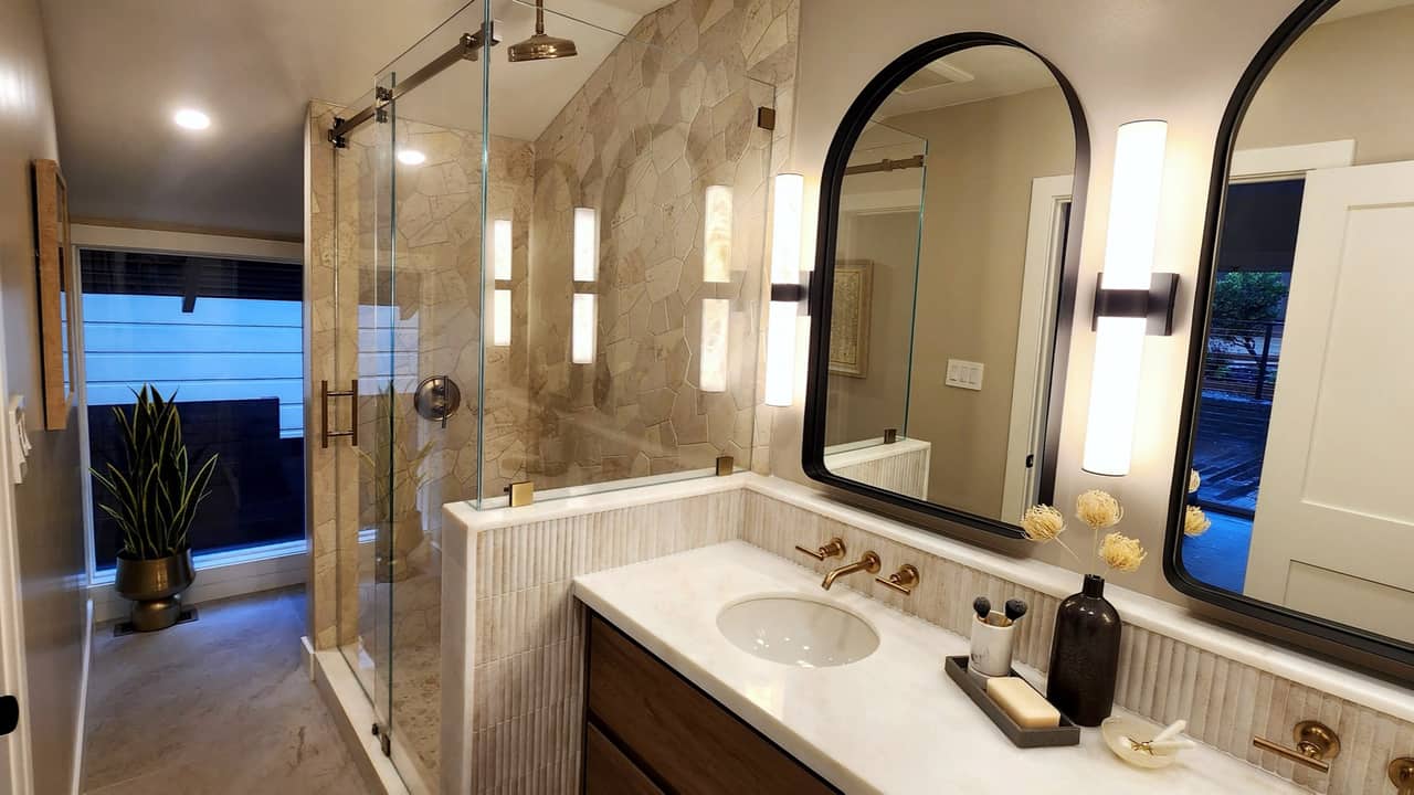 Frameless glass corner shower in beige stone tile with arched black mirrors and gold fixtures